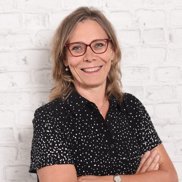 Frau mit blonden Haaren, Brille und gepunktetem Hemd, lächelt vor einer Backsteinmauer.